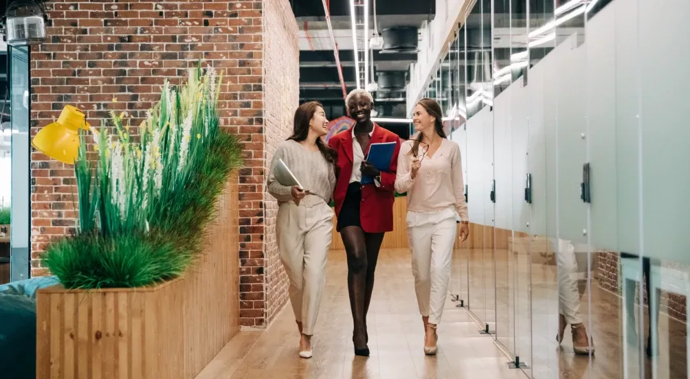 Tre personer går tillsammans genom en modern kontorskorridor, samtalar och ler, omgivna av glasväggar, gröna växter och tegelväggar.